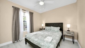 Ein Schlafzimmer mit Bett, Deckenventilator, Nachttisch mit Lampe und Fenster mit Vorhängen.