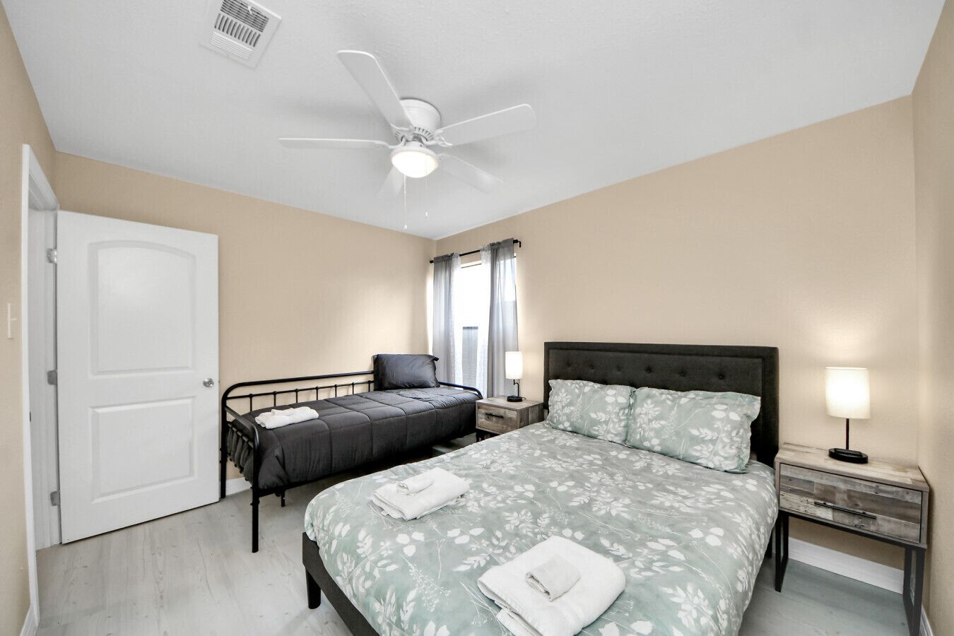 A bedroom with a bed, a smaller bed, a ceiling fan, a window with curtains, and a nightstand with a lamp.