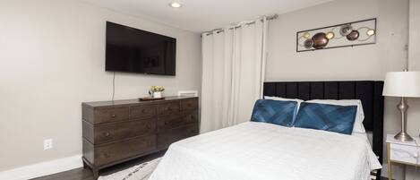 A bedroom with a bed, a dresser, a TV mounted on the wall, and a nightstand with a lamp.
