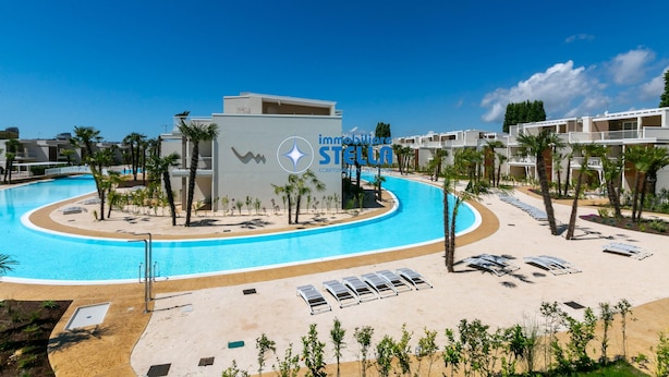 Ein Swimmingpool mit einem modernen Gebäude im Hintergrund, umgeben von Palmen und Liegestühlen.