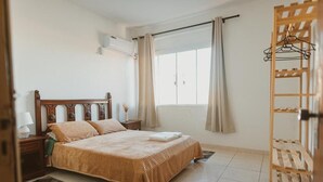 A bedroom with a bed, a nightstand, a lamp, a window with curtains, and a wooden rack.