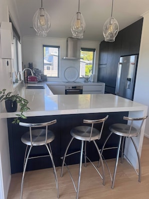 A modern kitchen with a central island, three bar stools, stainless steel appliances, and pendant lighting. - A Beautiful Beach Holiday Home in Pauanui (Pauanui)