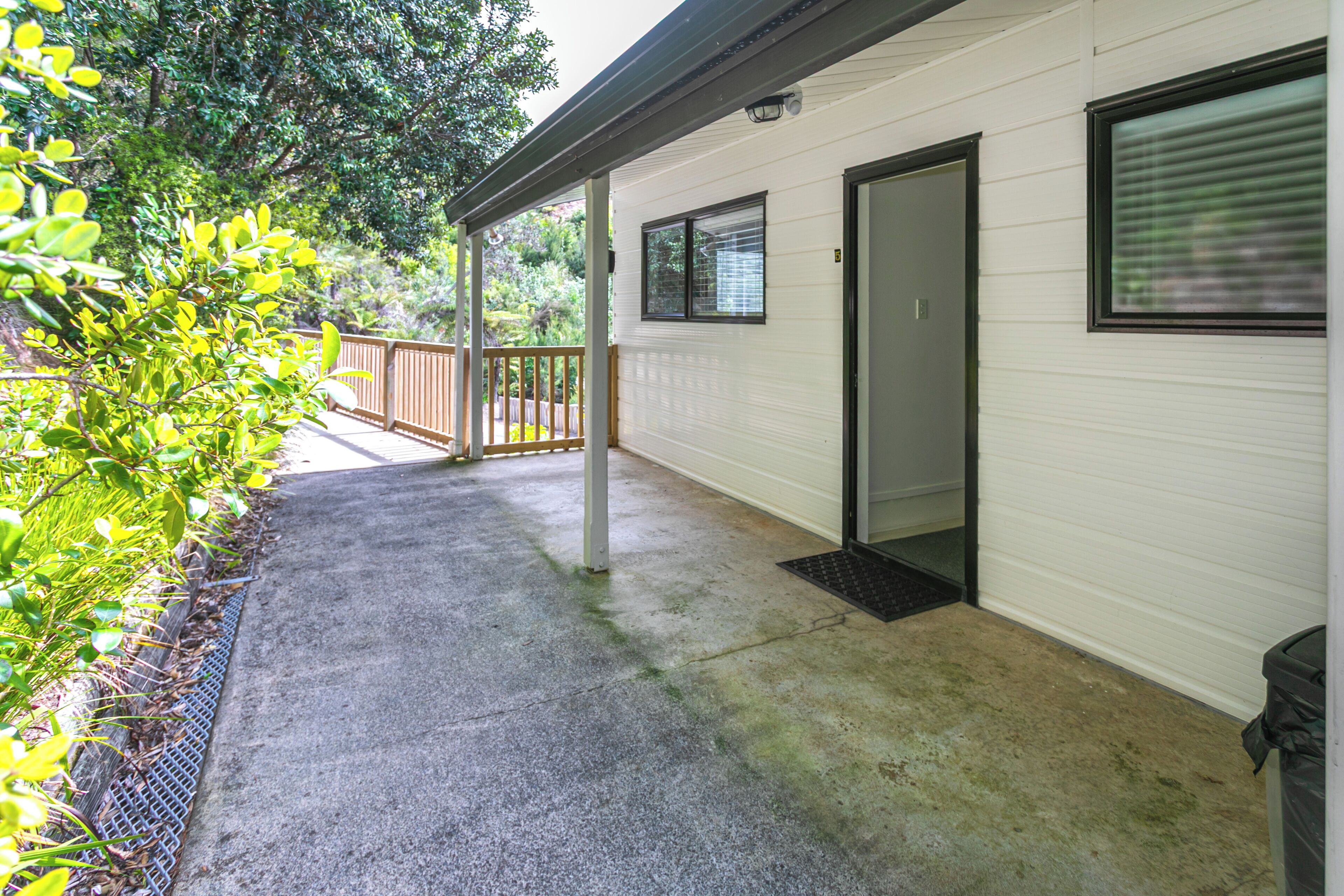 A covered outdoor area with a concrete floor, a wooden deck, and a white building with a door and windows.