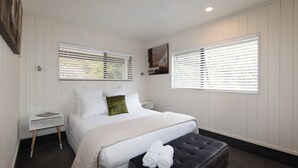 A bedroom with a bed, bedside tables, a window with blinds, and a dark-colored ottoman with towels. - Sweeping panoramic views of Hahei beach and offshore islands (Hahei)