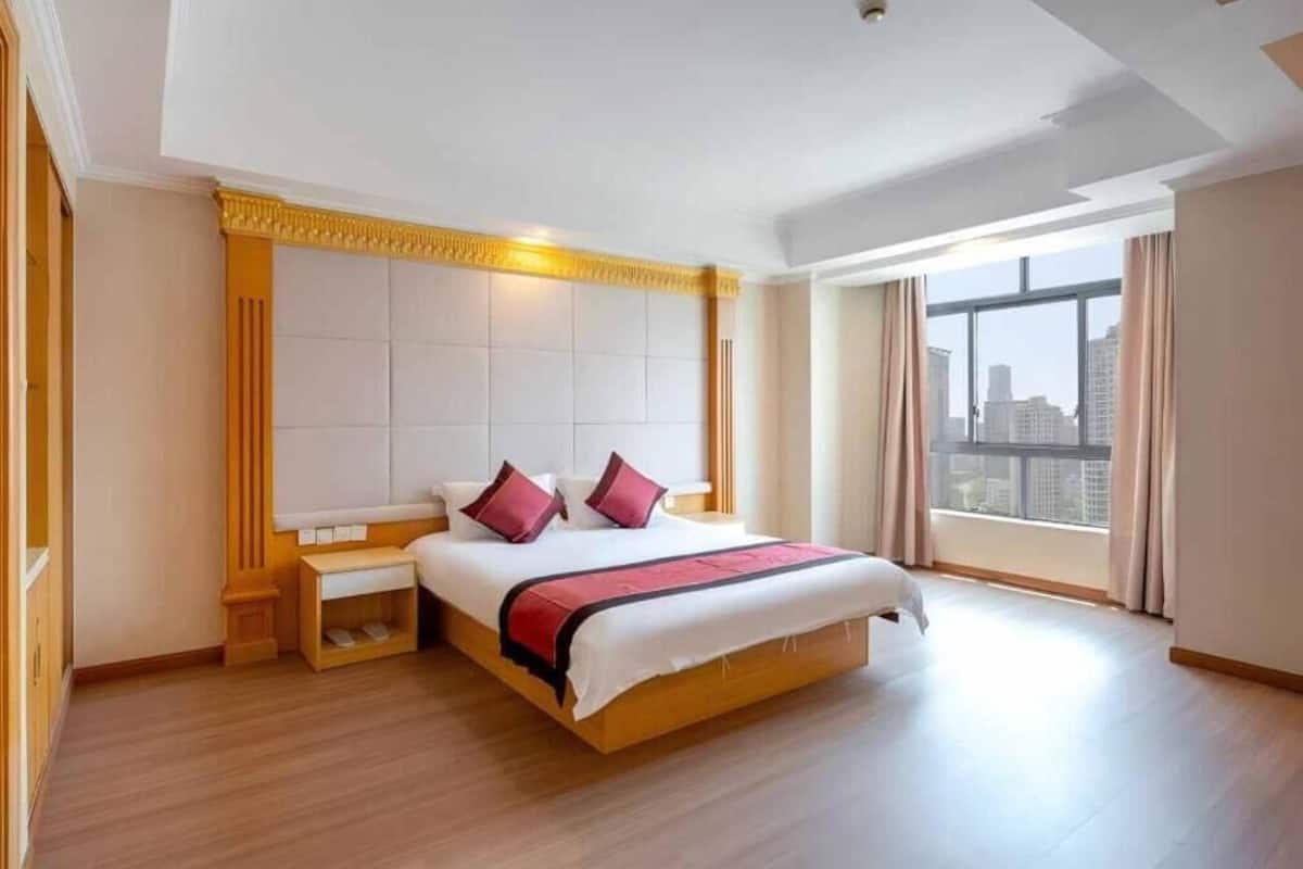 Une chambre d’hôtel avec un grand lit, une tête de lit en bois, des tables de chevet et une vue sur la ville grâce à de grandes fenêtres.