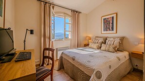 A bedroom with a bed, a desk with a computer, a chair, a window with curtains, a bedside lamp, and a wall-mounted picture. - GRIMAUD – BEAUTIFUL PROVENCAL VILLA IN THE HEART OF THE VILLAGE (GRIMAUD)