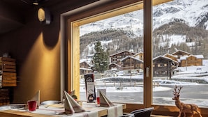 A dining area with a table set for two, a wooden interior, and a view of snow-covered buildings and mountains through large windows. - Cosy apartment for 6 guests with WIFI, TV and pets allowed (Zinal)