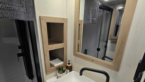 Casa de banho com uma pia branca, espelho, prateleira com artigos de higiene pessoal e zona de duche.