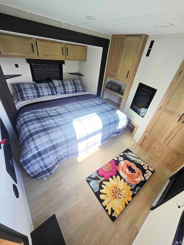 Dormitorio compacto de autocaravana con cama, un pequeño espacio de descanso, televisor integrado y armarios de madera.