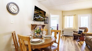 Een eetgedeelte met een houten tafel, stoelen, een klok en een aan de muur bevestigde televisie.