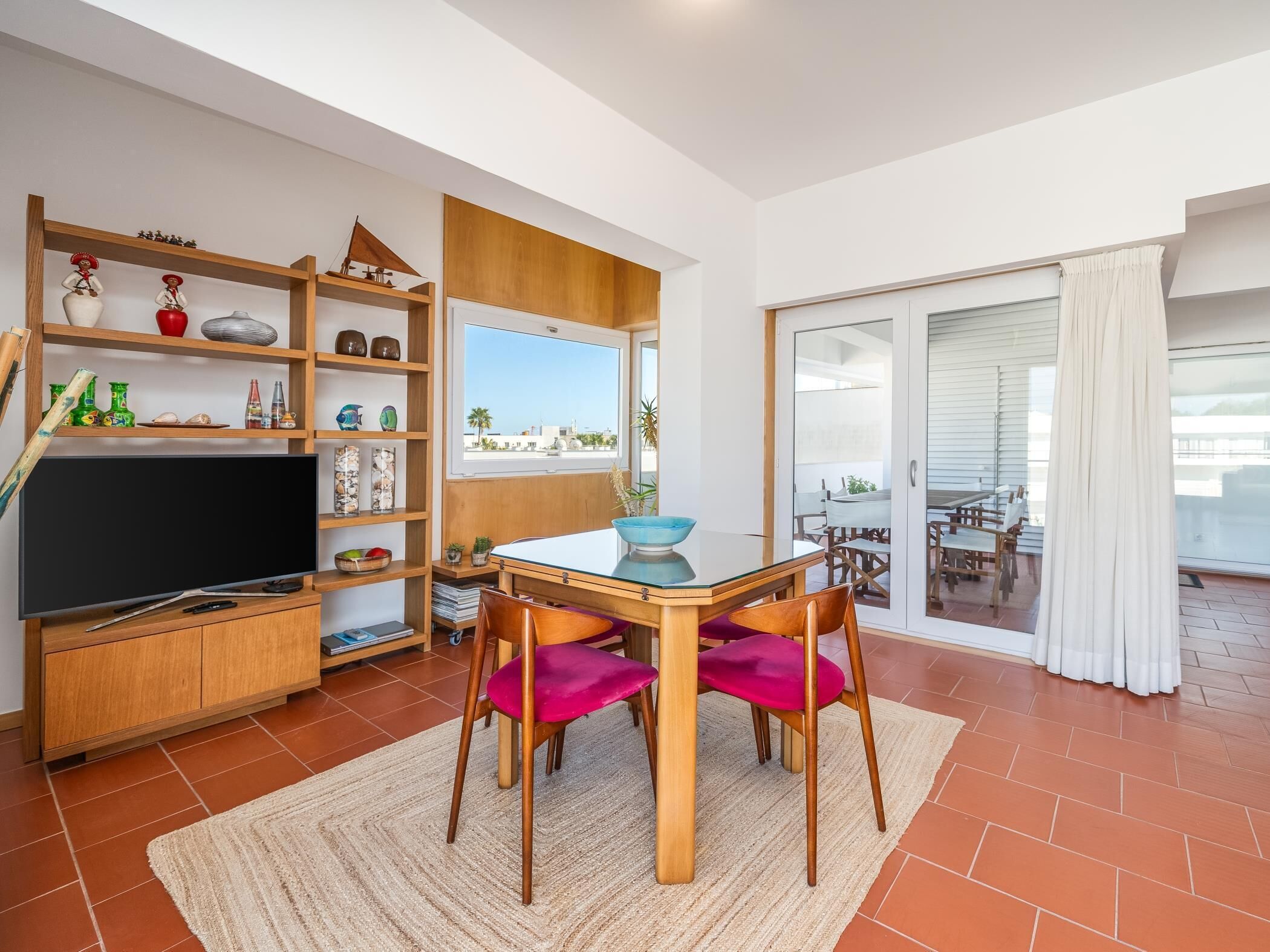 A living room with a TV, wooden shelves, a dining table with chairs, and a sliding glass door to a balcony.