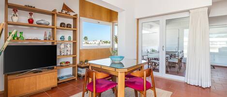 A living room with a TV, wooden shelves, a dining table with chairs, and a sliding glass door to a balcony.