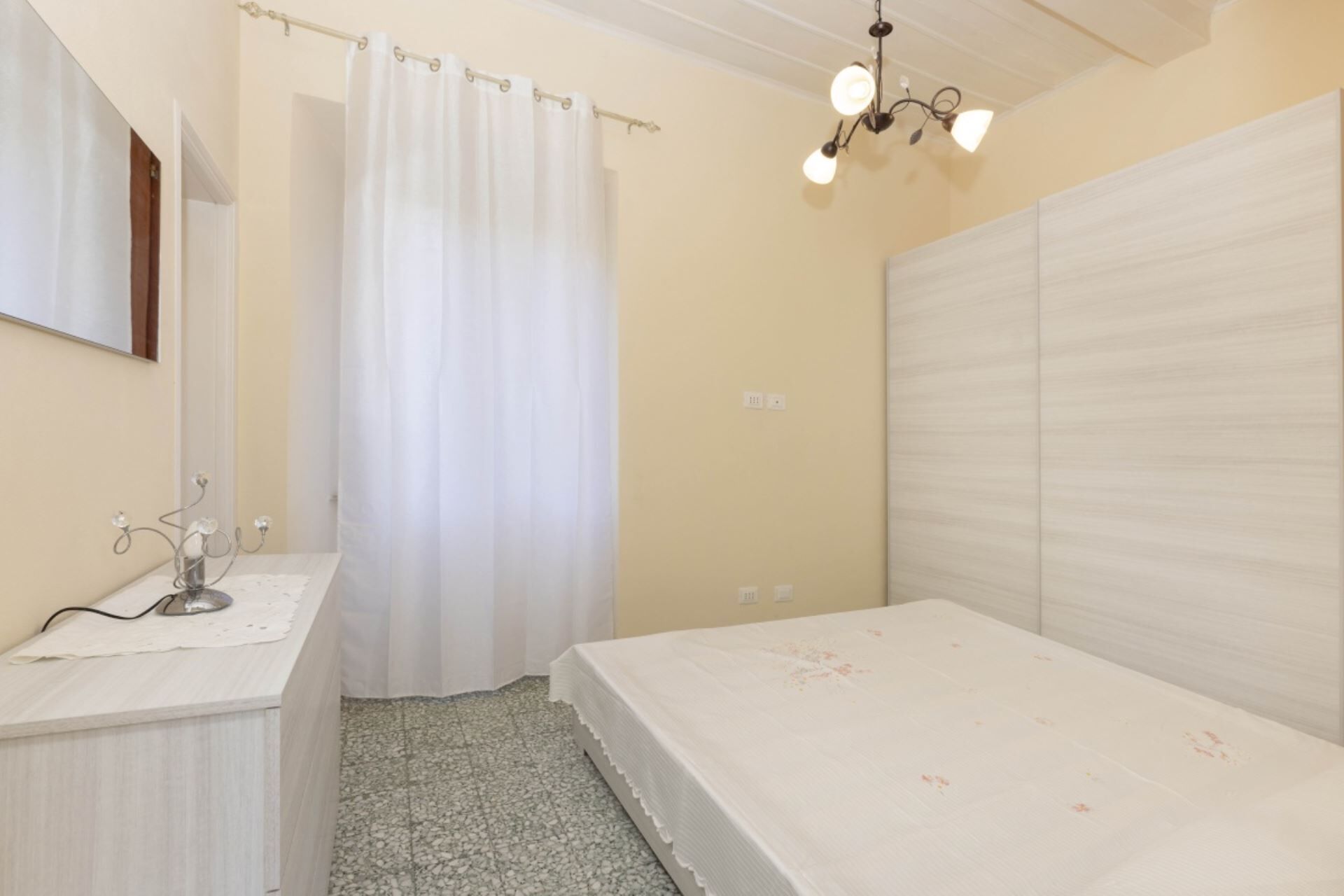 A bedroom with a bed, a bedside table, a mirror, a curtain, and a chandelier.