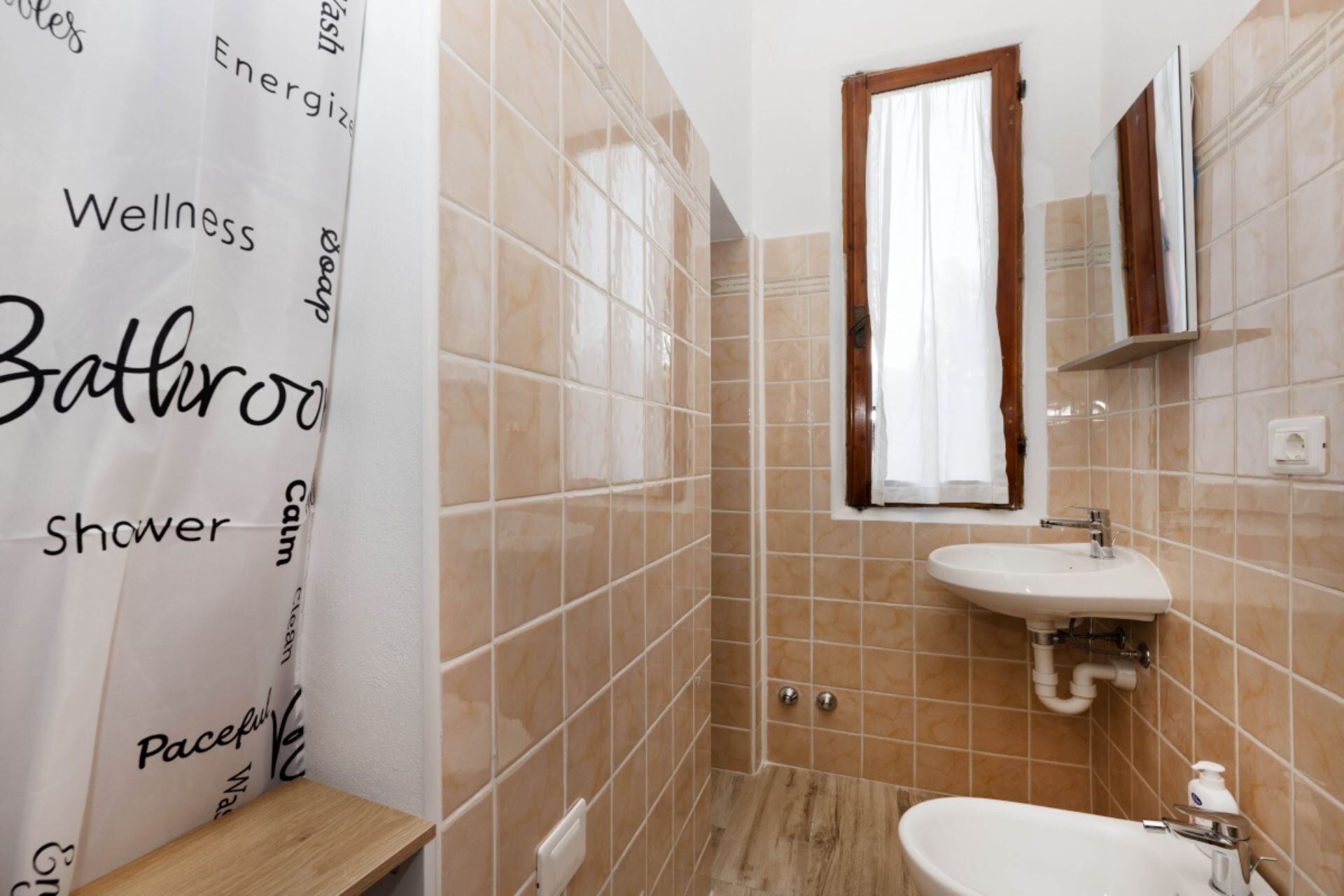 A bathroom with beige tiles, a white sink, a mirror, and a shower curtain with text.