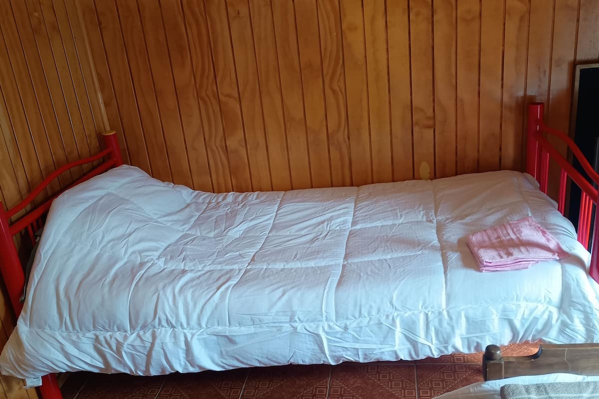 Una cama individual con edredón blanco y toalla rosa, paredes de paneles de madera y estructura metálica roja.