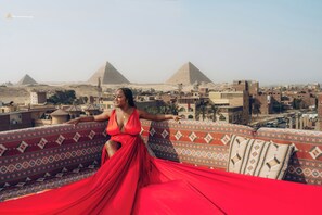 A person in a red dress sitting on a patterned sofa with a view of the pyramids in the background. - Solis pyramids view hotel (Giza)