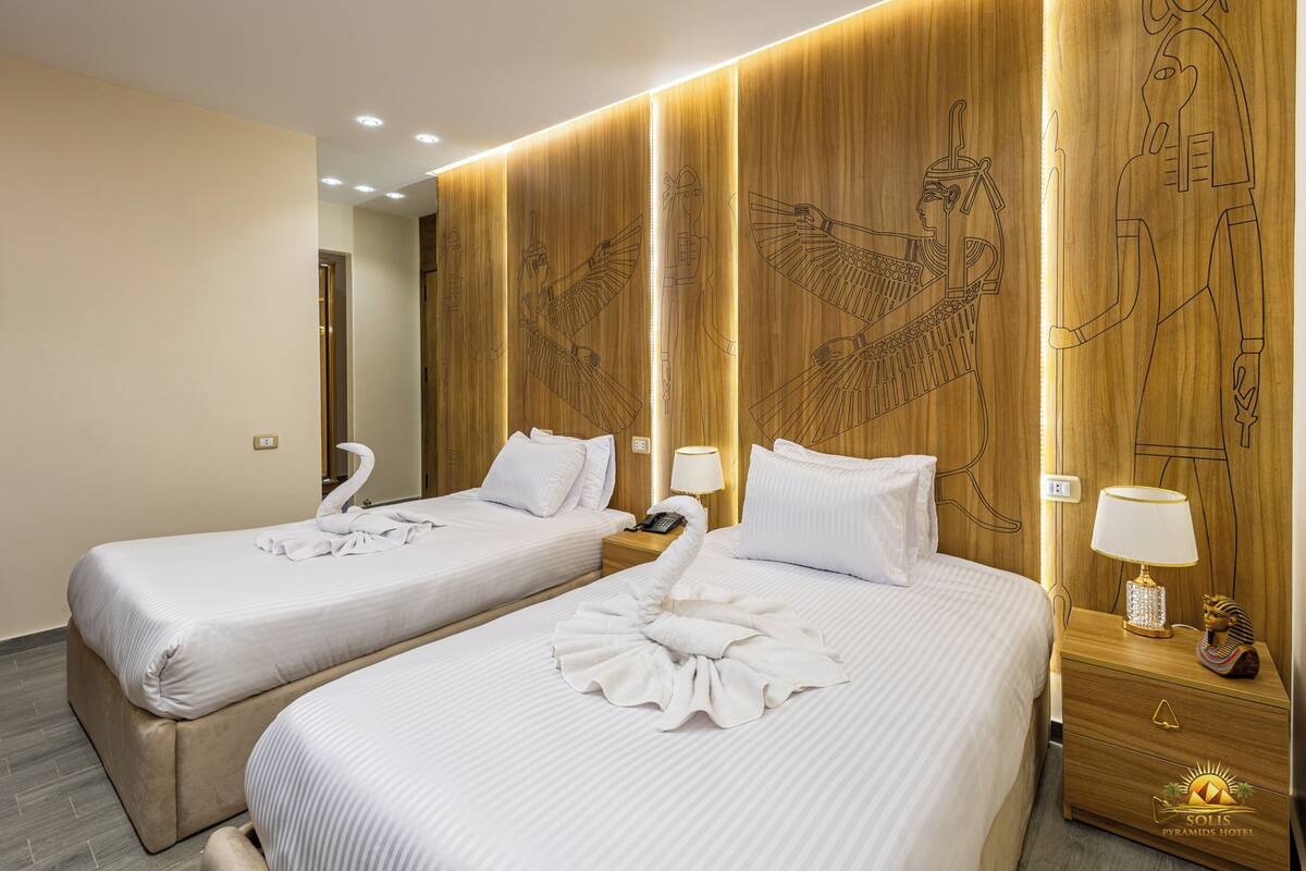 Ein Hotelzimmer mit zwei Betten, einem Holzkopfstück mit ägyptischen Hieroglyphen, weißen Bettwäsche und einem Nachttisch mit Lampe und Dekoration.