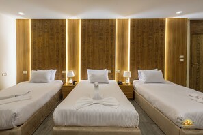 A hotel room with three single beds, wooden headboards with decorative carvings, bedside lamps, and a central bed with a towel arrangement.
