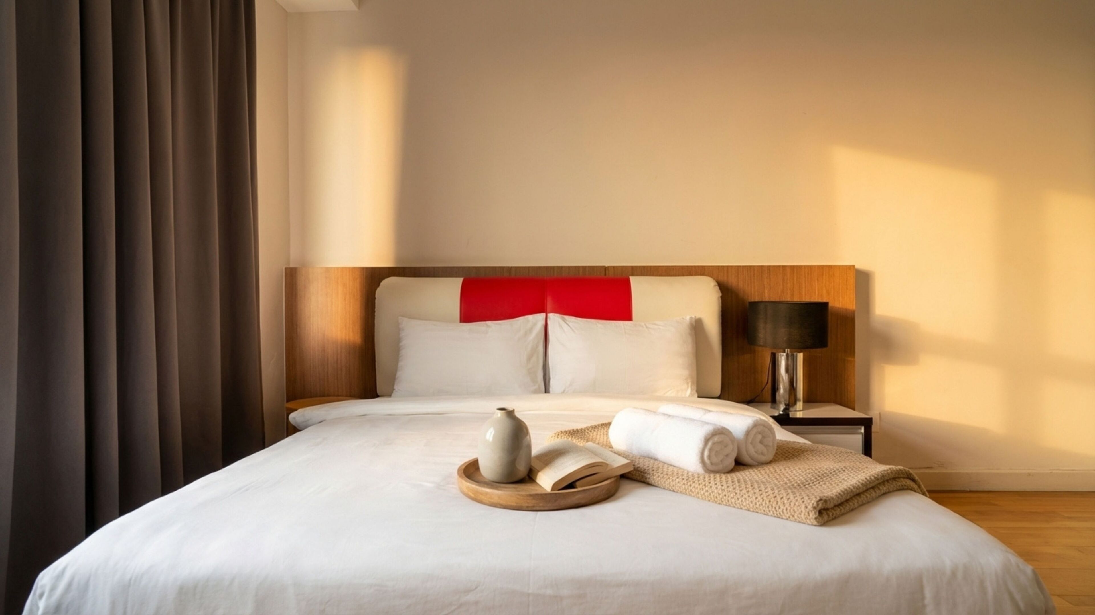 Una habitación de hotel con cama, mesita de noche con lámpara y ventana con cortinas.