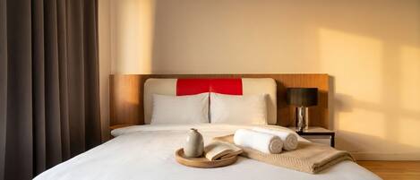 Una habitación de hotel con cama, mesita de noche con lámpara y ventana con cortinas.