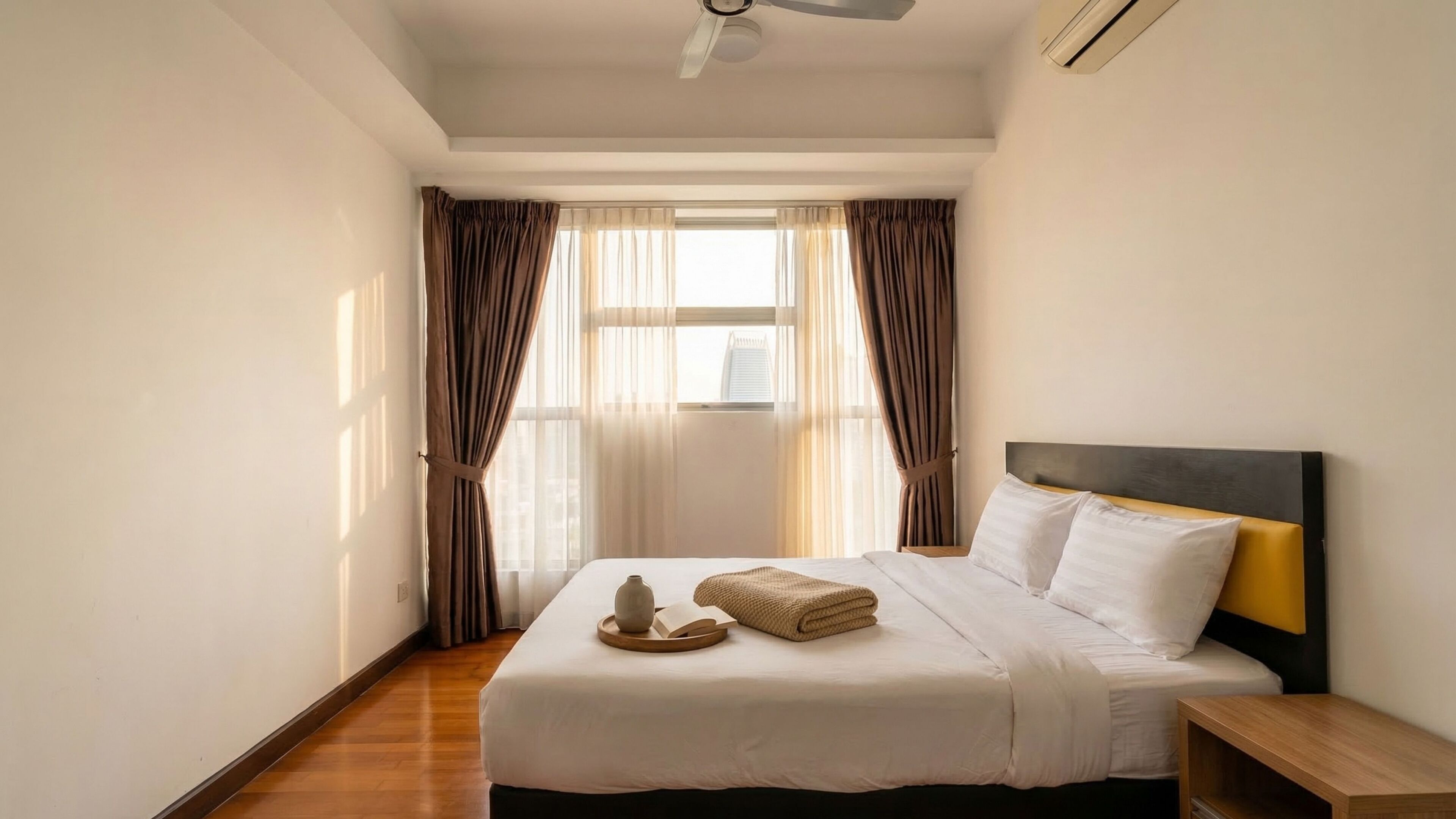 Hotellihuone, jossa on sänky, yöpöytä, ikkuna verhoilla ja kattotuuletin.