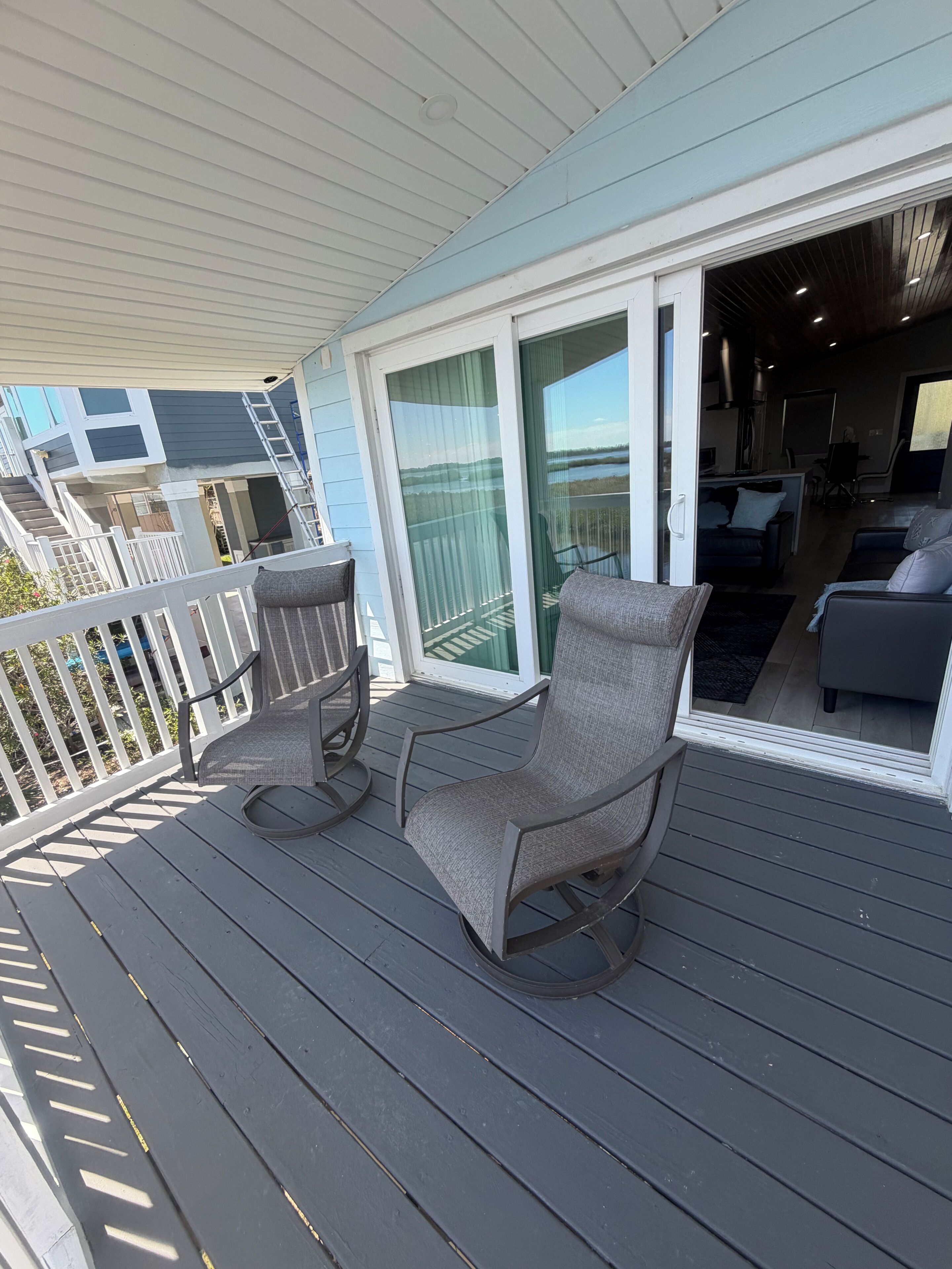 A covered deck with two chairs, a table, and a sliding glass door leading to an interior space with a sofa and a dining area.