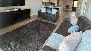 Ein modernes Wohnzimmer mit Holzboden, einem großen Flachbildfernseher, einer Couch, einem Couchtisch und einer Küche im Hintergrund.