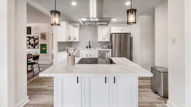 Cocina moderna con armarios blancos, una isla central, electrodomésticos de acero inoxidable e iluminación colgante.