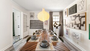 Comedor con una larga mesa de madera dispuesta para seis personas, una lámpara colgante y varias obras de arte en las paredes.