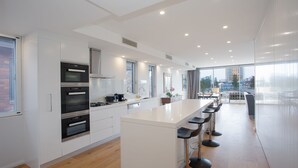 A modern kitchen with built-in ovens, a central island with bar stools, and a large window overlooking a cityscape. - Saltview, Queenscliff (Queenscliff)