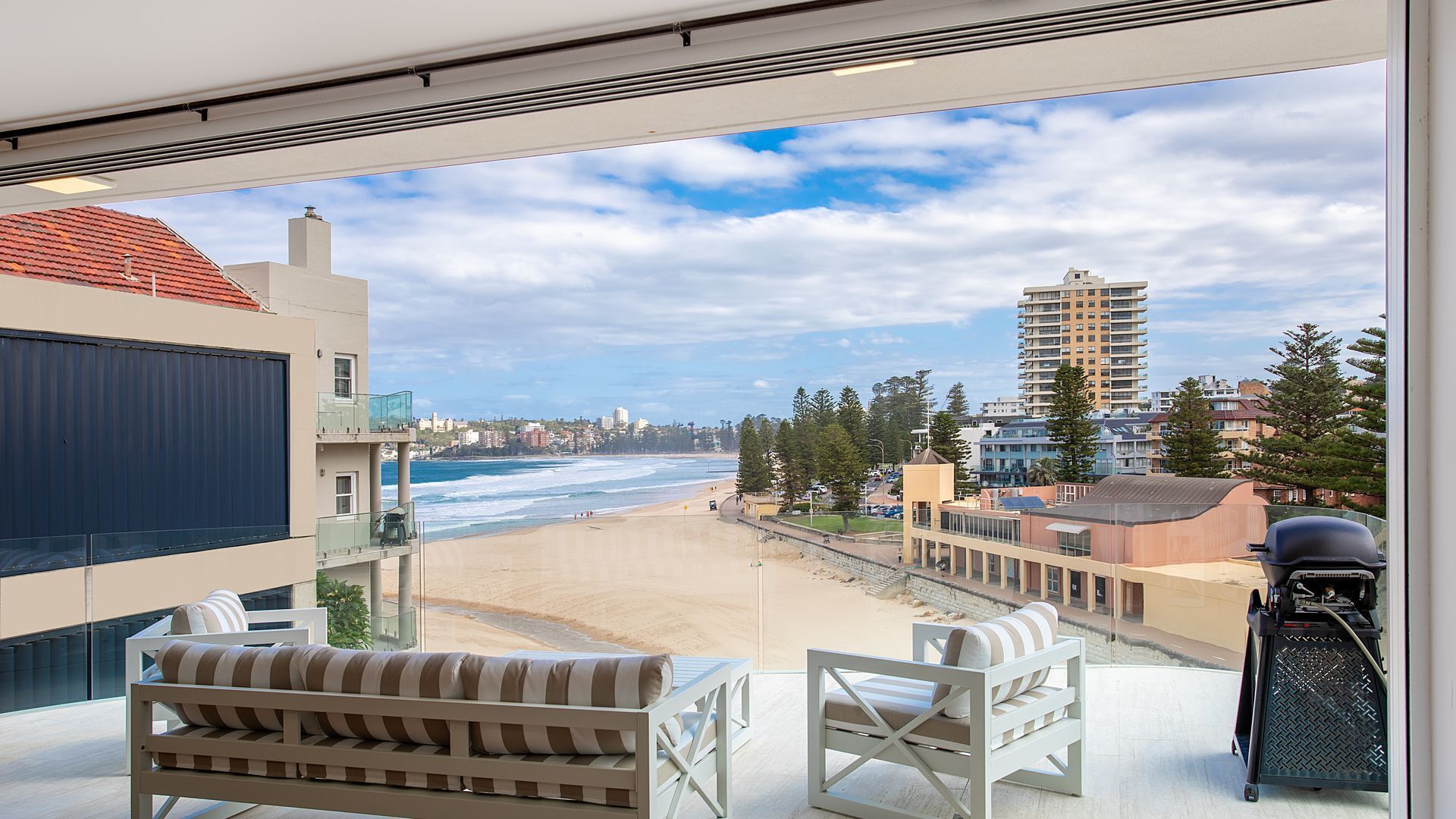 A balcony with a view of the beach, a barbecue grill, and a seating area with cushions.