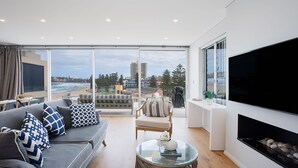 A modern living room with a large TV, a sofa with patterned cushions, a glass coffee table, and a view of the beach and buildings. - Saltview, Queenscliff (Queenscliff)