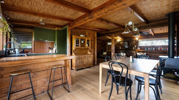 A rustic cabin interior with a wooden dining table, bar stools, a fireplace, and a bookshelf. - Wattlewood  | Mid Century Chic with Outdoor Serenity (Frankston South)