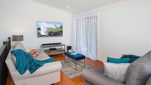 A living room with a sofa, a coffee table, a TV, and a window with sheer curtains. - 4BR Pet friendly home with ocean views (Kiama)