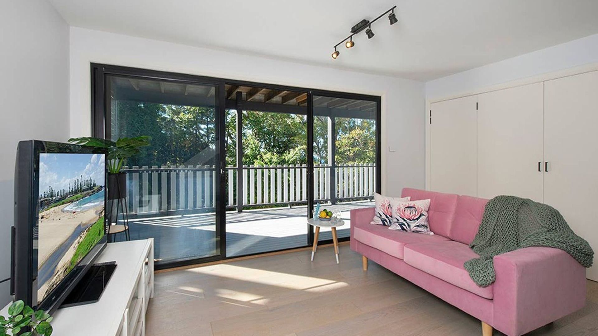 A modern living room with a pink sofa, a television, and a balcony with a view of the beach.