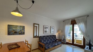 Ett vardagsrum med en soffa, ett matbord, stolar, en lampa och en fläkt. På väggen hänger inramade tavlor och det finns ett fönster med gardiner.