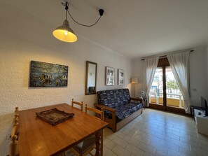 A living room with a wooden dining table, a patterned sofa, a ceiling light, and a window with curtains. - Apartment 1 minute from Riells Beach (l'Escala)