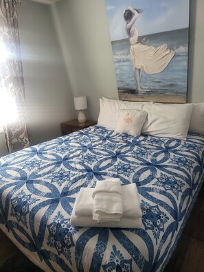 A bedroom with a patterned bedspread, pillows, a folded towel, a painting of a woman by the sea, and a nightstand with a lamp.