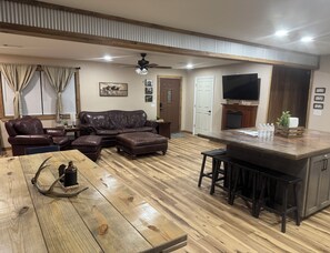 A living room with a sofa, ottoman, wooden coffee table, and a kitchen area with a bar stool and countertop.