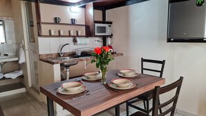 Un coin repas avec une table en bois dressée pour quatre personnes, un vase rempli de roses rouges et, au fond, une cuisine équipée d’un four à micro-ondes et de tasses à café.