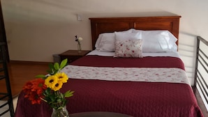 Un lit avec une tête de lit en bois, une couvre-lit rouge et un coussin à motifs floraux. Il y a une table de chevet avec un vase de fleurs.