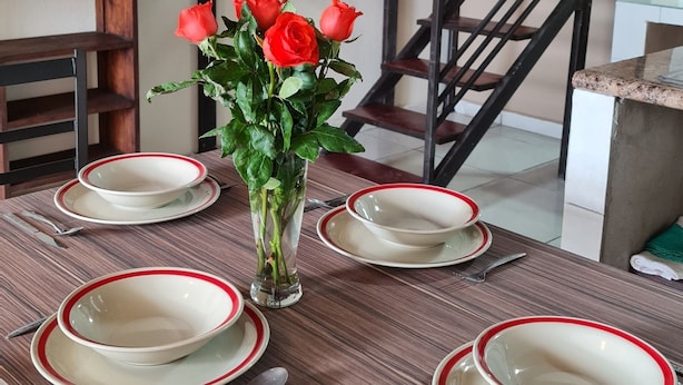 Une table à manger dressée avec des assiettes et des couverts, un vase rempli de roses rouges et un escalier en arrière-plan.