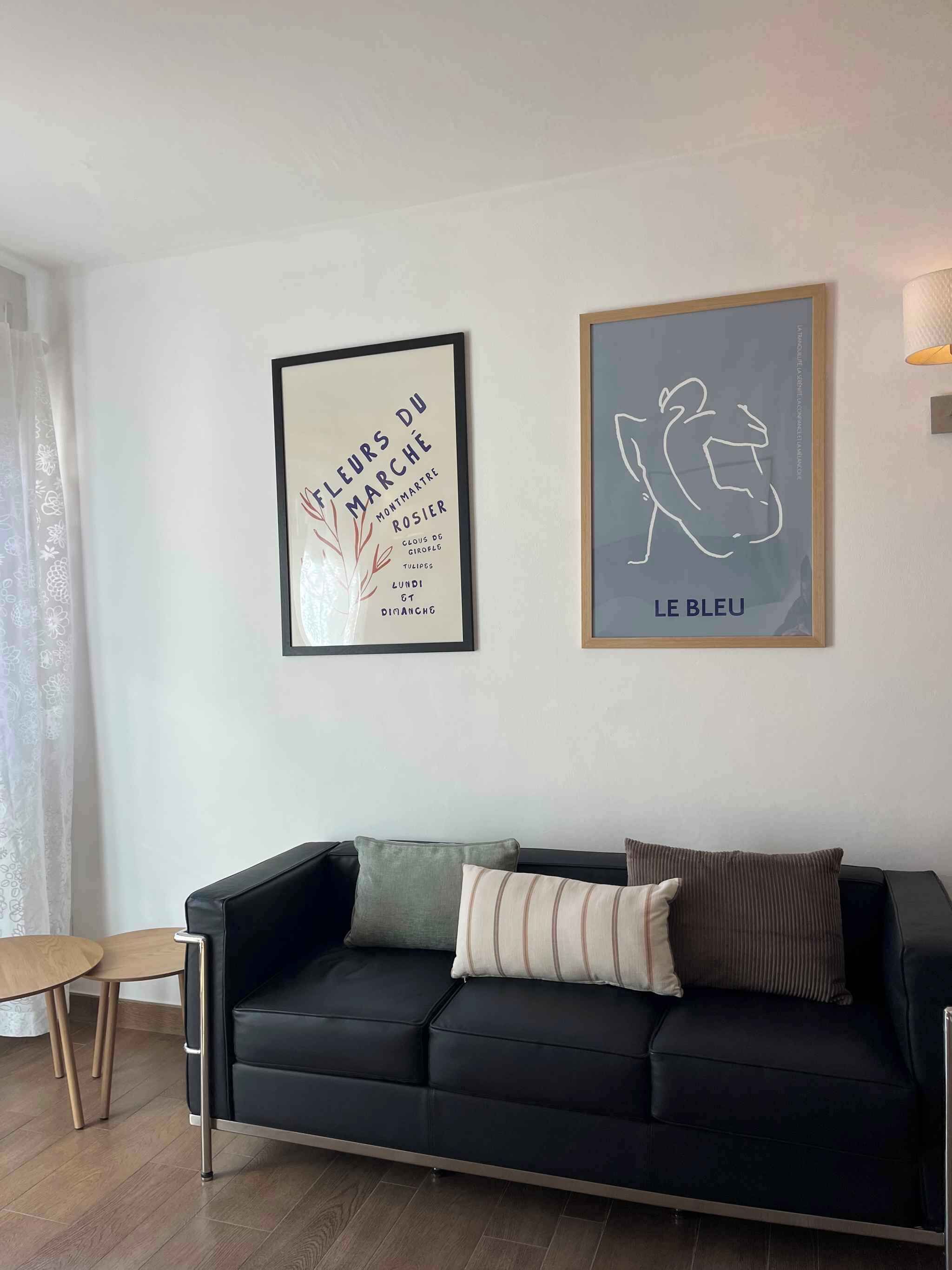 Un salon moderne avec un canapé en cuir noir, une table d’appoint en bois et deux œuvres d’art encadrées accrochées au mur.