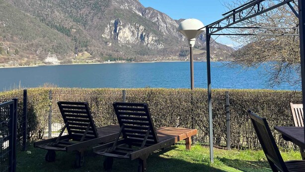 Eine Terrasse mit Liegestühlen, einem Tisch und einer Lampe mit Blick auf einen See und Berge.