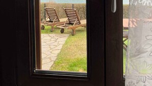 Blick durch ein Fenster mit Türgriff, zwei Lounge-Sessel auf einer Steinterrasse und eine gebirgige Landschaft im Hintergrund.