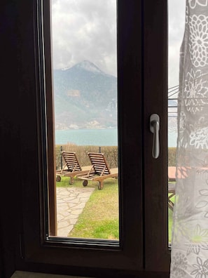 Blick durch ein Fenster mit Türgriff, zwei Lounge-Sessel auf einer Steinterrasse und eine gebirgige Landschaft im Hintergrund.