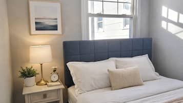 Ein Schlafzimmer mit Bett, Nachttisch, Lampe, Uhr, gerahmtem Bild, Fenster und einer Deckenleuchte.