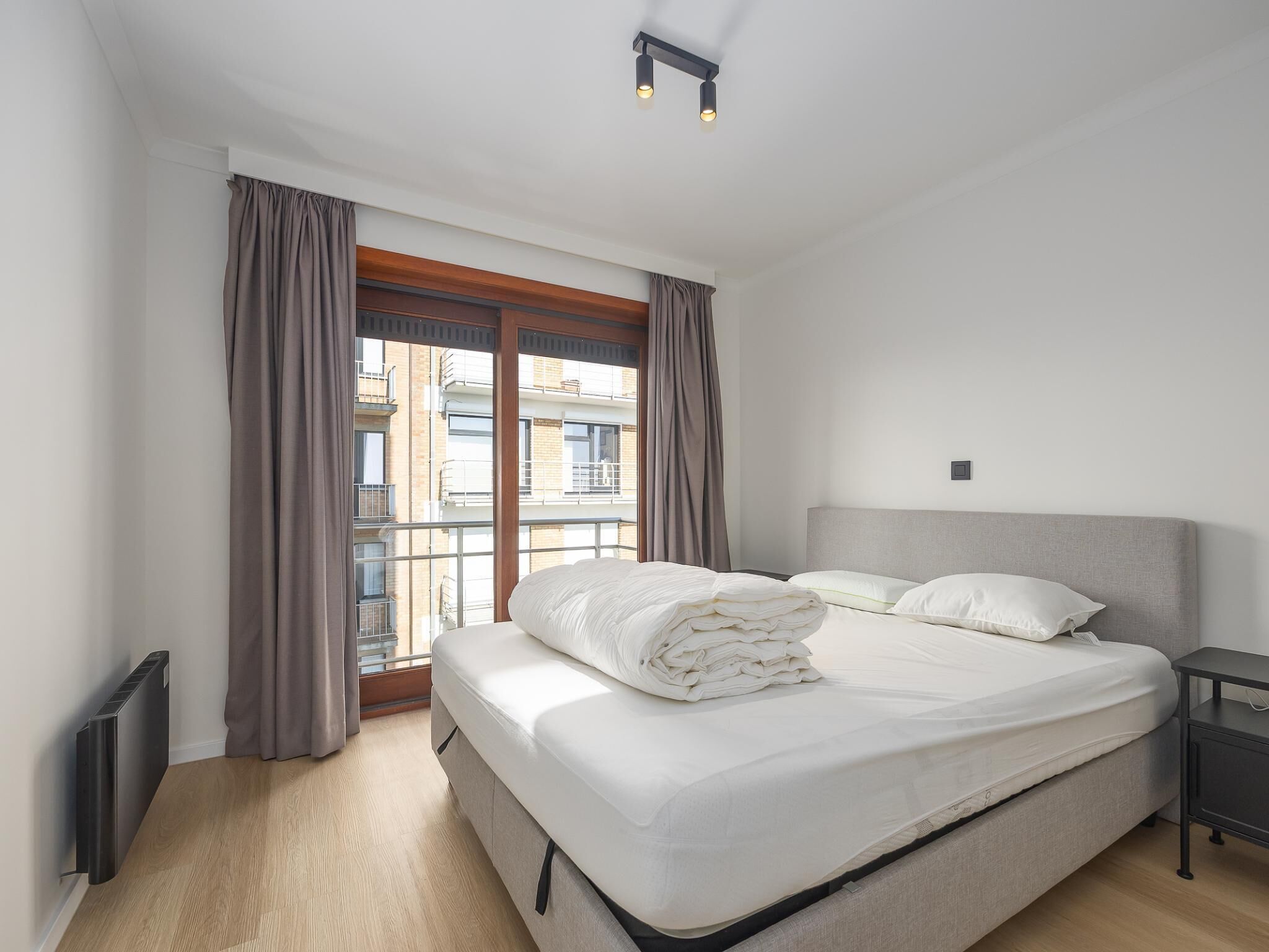 A bedroom with a bed, a nightstand, a window with curtains, and a radiator.
