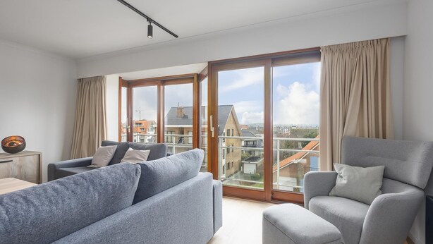 Ein modernes Wohnzimmer mit einer Couch, einem Sessel und Blick auf Gebäude durch große Fenster.