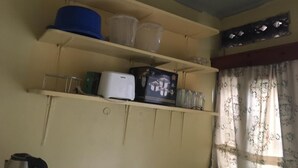 Een kamer met beige wanden, een plafond met een rasterpatroon, en een raam met bloemen gordijnen. Er staan planken met diverse spullen, waaronder een blauwe hoed, plastic bakken en een wit apparaat.